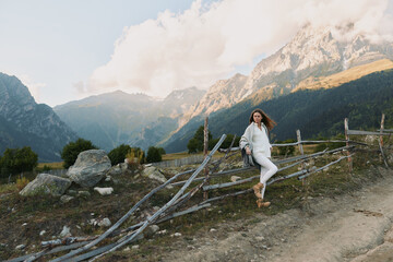 Fototapeta premium Young woman in white outfit leans on rustic wooden fence with mountain landscape in the background during a clear day.