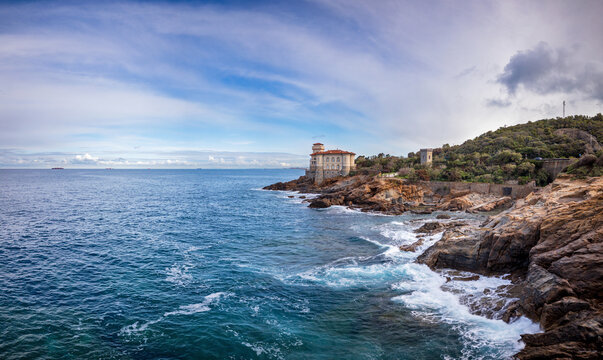 View of Castello del Boccale perched on a rocky cliff overlooking the blue Mediterranean sea with crashing waves and green hills in Calafuria, Toscana, Italy.