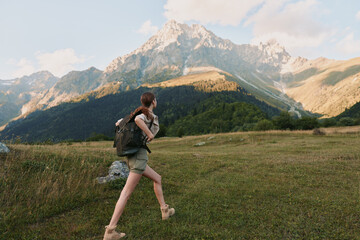 Fototapeta premium A woman with a backpack hikes through a grassy meadow towards tall mountains under a partly cloudy sky. The scene captures outdoor adventure and lifestyle in nature.