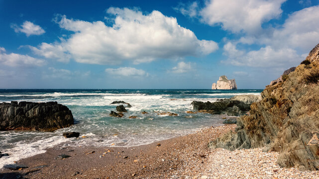 View of Scoglio Pan di zucchero sea stack from the rocky shore of Masua beach with crashing waves and white clouds in a blue sky in Masua, Sardegna, Italy.