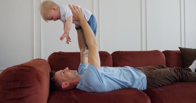 Happy father lying on a rust colored sofa lifts and cuddles his baby in a bright living room, sharing playful bonding time at home