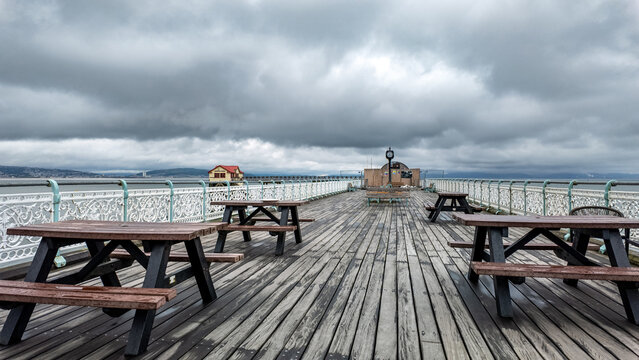 View of Mumbles Pier featuring wooden picnic tables and ornate white railings under a dramatic overcast sky in Mumbles, Wales, United Kingdom.