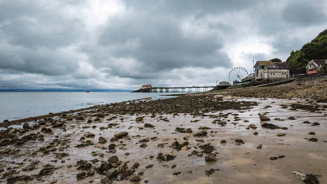 View of Mumbles Pier stretching into the sea with a Ferris wheel and rocky beach under a cloudy sky Mumbles, Wales, United Kingdom.