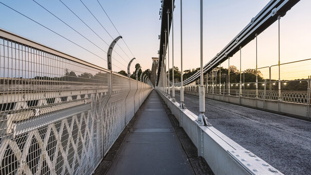 View of Clifton Suspension Bridge showing the pedestrian walkway with metal fencing and suspension cables under a clear sky at dusk in Bristol, United Kingdom.