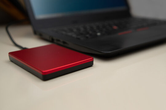 Angled shot of a red portable external memory drive connected to a modern laptop on a desk. Premium hardware solution for personal and business data storage.