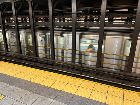 View of a New York City subway train moving past a station platform with yellow tactile paving and dark structural pillars New York, New York, United States.