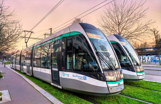 Saint Denis, France - December 29, 2025: Two modern Alstom passenger trams on green grass tracks at sunset. The public transport vehicles operate near Paris.