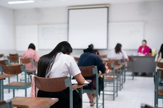 Rear view of female student in uniform taking test in university classroom with teacher in pink jacket at desk, authentic academic exam evaluation.