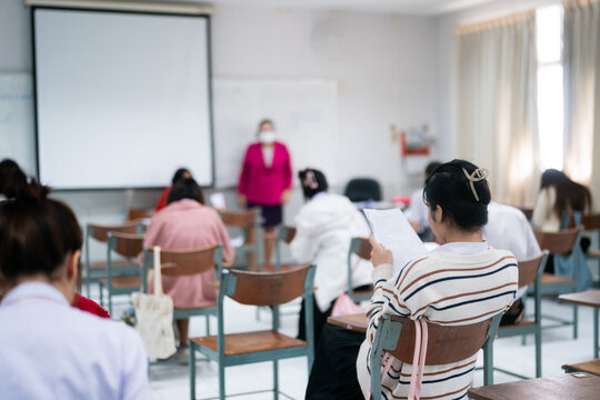 Female student holding test paper during exam with proctor standing in blurred university classroom, authentic academic evaluation, rear view.