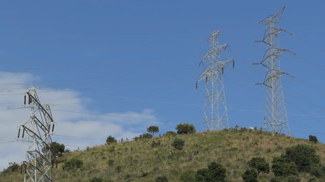 High-voltage electricity pylons on a green hill with clouds moving in the blue sky, representing power transmission, energy grid, and industrial infrastructure