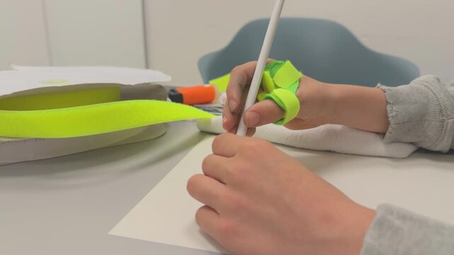 Child with dysgraphia using an ergonomic pencil grip during occupational therapy session while an adult therapist is assisting with hand positioning and fine motor skills