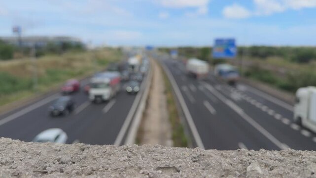 Busy highway traffic with blurred cars and trucks seen from an overpass with concrete wall in the foreground creating a cinematic bokeh effect on a sunny day