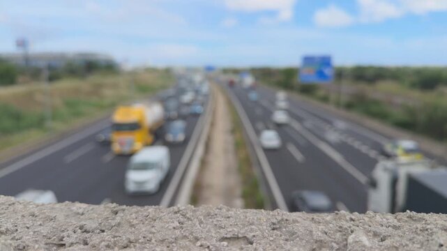 Concrete overpass barrier with a blurry background of highway traffic, showcasing vehicles like cars and trucks driving along the road under a blue sky during the daytime