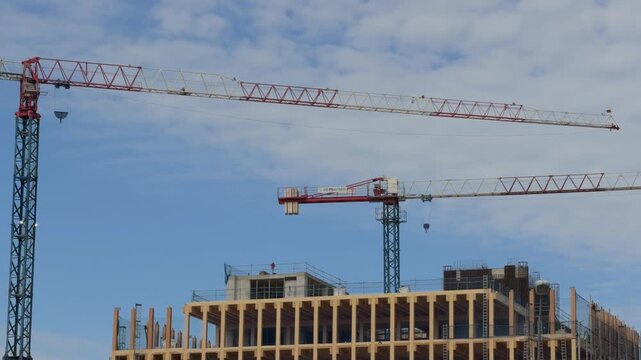 Construction site featuring towering cranes lifting materials over a modern wooden and concrete building structure, showcasing architectural progress under a blue sky with clouds