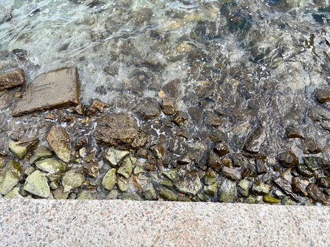 A high-angle shot of crystal clear ocean water flowing over smooth sea stones and pebbles, creating natural textures and foam on a sunny day.