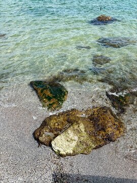 A top-view shot of transparent turquoise ocean water gently rippling over a sandy shore with natural stones and green moss under bright sunlight