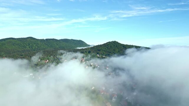 A serene, forested landscape with a small village nestled among lush greenery, partially obscured by misty clouds, showcasing tranquil natural beauty and peaceful coexistence of human habitation.