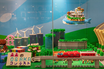 Obraz premium A scene made of building blocks shows a vibrant city with a flying ship, wind turbines, parks, and various small figures. This display captures a lively urban environment