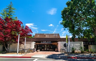 Fototapeta premium Palo Alto, California - May 29, 2023: Exterior view of the Palo Alto City Library Downtown Branch building, featuring bright red and green foliage against a blue sky in Silicon Valley, United States