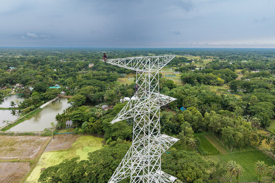 Trishal Upazila, Bangladesh - 13 May 2026: Aerial view of workers on a tall white electricity pylon surrounded by lush green fields and ponds under an overcast sky.