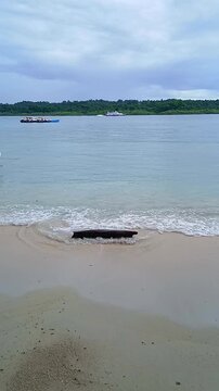 Calm tropical beach with waves washing over a log, distant boats, and a cloudy sky