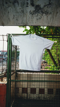 White t-shirt hangs on a metal fence. Green leaves frame the top of the shot. Background shows distant buildings and trees. Soft daylight casts gentle shadows. Image suggests a quiet, everyday moment