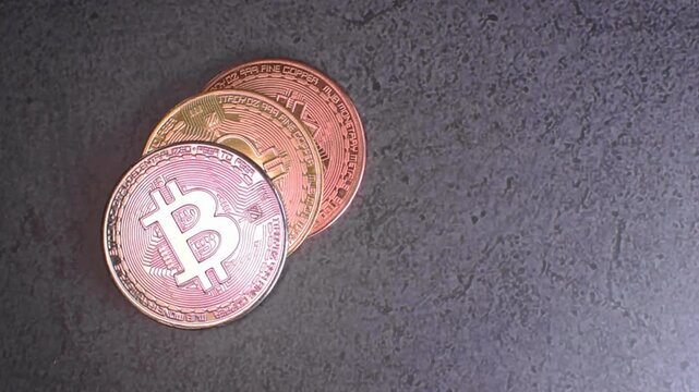 Three bitcoin cryptocurrency coins stacked on a gray granite surface with copper, gold, and silver finishes facing the camera while moving light creates glowing reflections and metallic highlights, re