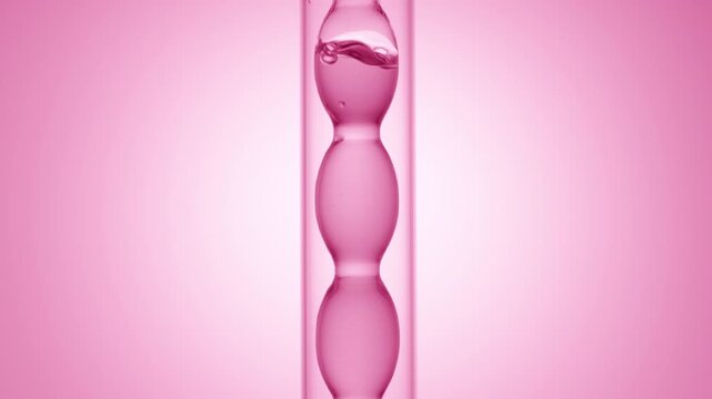Tight zoom on air bubbles bursting through glossy clear pink fluid as it fills and drains from a bulb laboratory condenser on pink ramp background