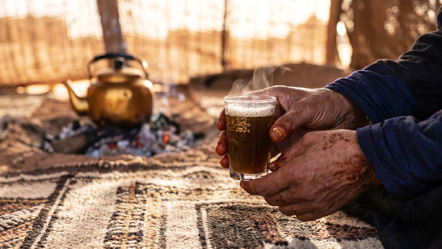 Tuareg Artisan Henna-Darkened Hands Holding Ornate Glass of Saharan Mint Tea on Woven Rug in Desert Tent Near Agadez Niger, North African Culture, Nomadic Tradition