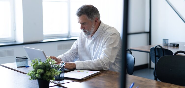 Smiling mature business man executive wearing shirt sitting at desk using laptop. Happy busy professional middle aged businessman investor working on computer looking away in office