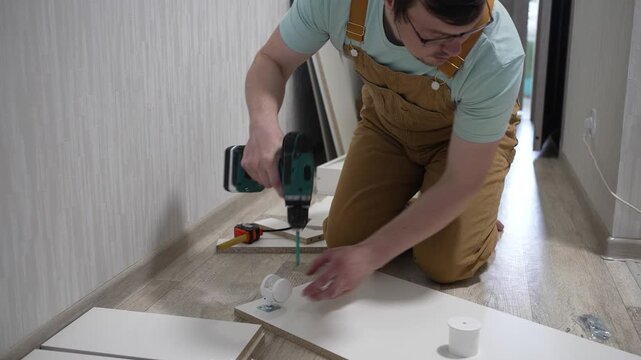 Focused young craftsman wearing overalls and glasses using a cordless electric drill to assemble flat-pack furniture, meticulously installing components on the floor of a new apartment