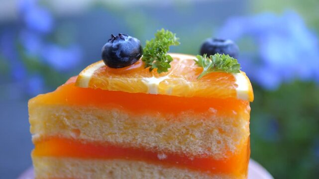 Cinematic close-up of a delicious slice of layered orange sponge cake topped with fresh orange slice and blueberries, blurred blue flowers background