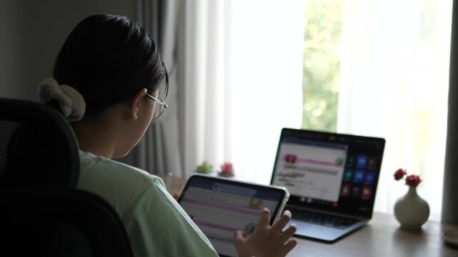 Cinematic wide over the shoulder shot of a young female professional with glasses holding a digital tablet and working on laptop at bright desk setup