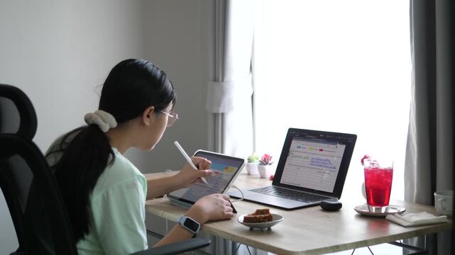 Cinematic wide over-the-shoulder shot of a young female creative professional using a stylus on digital tablet, multitasking with laptop on desk