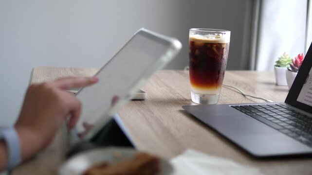 Cinematic medium shot of a professional using a digital tablet on a wooden desk, multi-tasking workspace with laptop and iced layered coffee background