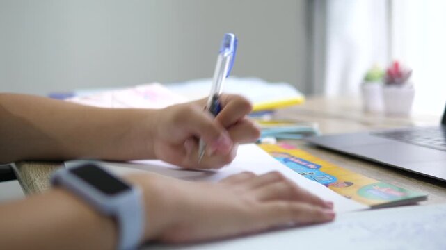 Cinematic close-up of a student's hand holding a pen and writing on a notebook, children studying and doing homework on desk with laptop background