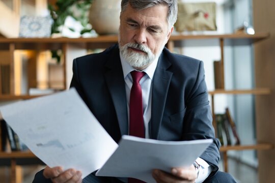 Mature businessman in formal suit concentrating on reading the report, sitting in the lobby and preparing for the meeting.