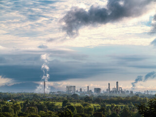 Obraz premium Distant view of a heavy industrial complex releasing steam, surrounded by lush green forest under a cloudy morning sky