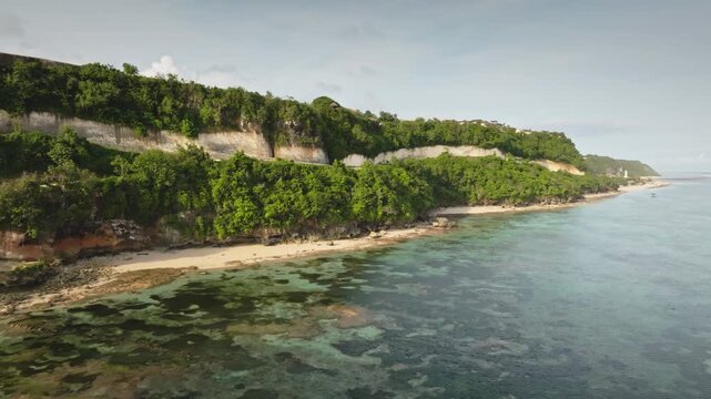 Tropical Green Bowl Beach on Bali's Bukit Peninsula with clear turquoise ocean revealing underwater reefs and a lush green cliffside creating a serene coastal landscape. Aerial view drone panorama