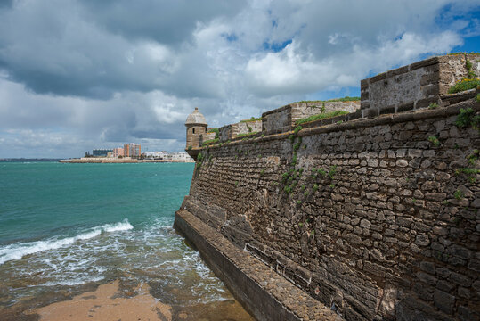 Detail of the defensive walls of Castillo de San Sebastian in Cadiz, Spain, with rough stone textures, Atlantic waves and dramatic cloudy sky along the historic coastal fortifications.
