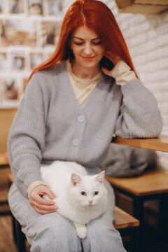 A red haired lady sits comfortably in a cozy cat cafe, gently holding a white cat on her knees while enjoying a peaceful warm relaxing indoor atmosphere soft natural light.