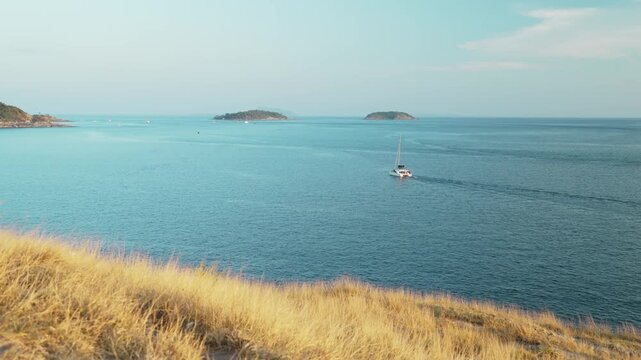 Serene Coastal Landscape Sailboat Gliding Across Calm Blue Sea, Surrounded by Distant Islands and Golden Grass in Foreground