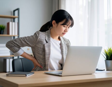 明るい室内でノートパソコンでの作業中に腰の痛みを感じ、辛そうな表情で腰に手を当てるジャケット姿の女性。健康・ヘルスケア