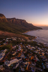Aerial view of Cape Town at sunset shows the Twelve Apostles above Camps Bay, a curving beach and rocky shore, villas and winding roads with warm lights, and a purple ocean. © Aerial Film Studio