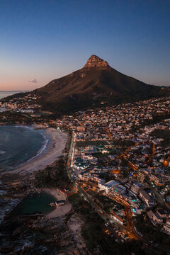 Aerial dusk view of Cape Town, South Africa, with Lion's Head rising above the Atlantic shore. Streetlights trace curving roads and beach as the sky shifts to purples.