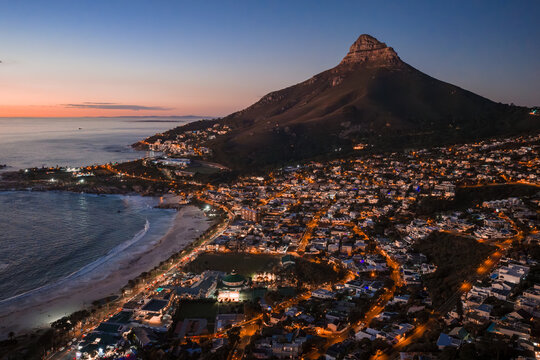 Aerial dusk scene in Cape Town shows Lion's Head above Camps Bay and Clifton. Streetlights trace winding roads and surf lines crescent beaches under purple orange twilight.