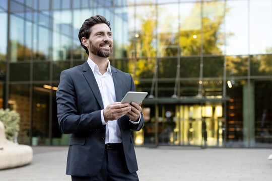 Confident businessman in a suit holding a digital tablet and smiling, standing outdoors in front of a modern glass office building, representing corporate success and technology-driven communication
