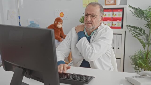 Male doctor at computer rubbing his shoulder and wincing while charting at desk in pediatric exam building with toys and files; fatigue strain ergonomic discomfort.