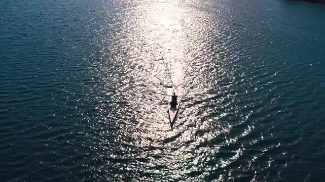 Drone shot of rowers moving across sunlit lake water with bright reflections and ripples. Peaceful aerial scene showing outdoor sport, endurance, teamwork, freedom and nature.
