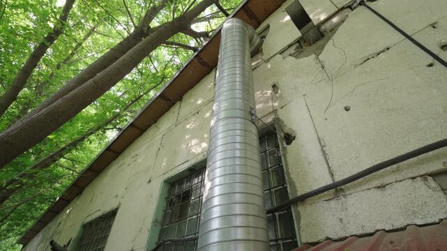 Low-Angle Bottom-Up View: Metal Ventilation Pipe on Old Industrial Building Exterior Wall | Galvanized Steel Chimney Duct Against Green Trees and Sky | Urban Infrastructure Detail